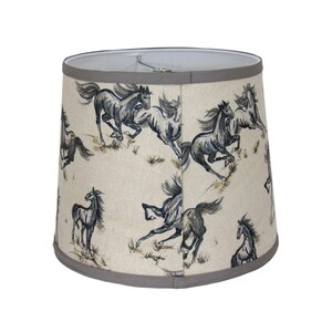 Op de afbeelding: Een beige lampenkap met een herhaald patroon van rennende paarden in grijstinten en zwart. De paarden zijn afgebeeld in een dynamische stijl, tegen een achtergrond van schaars gras. De kap heeft een grijze rand.