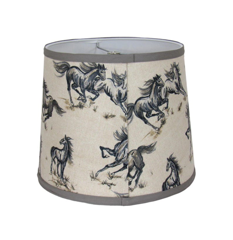 Op de afbeelding: Een beige lampenkap met een herhaald patroon van rennende paarden in grijstinten en zwart. De paarden zijn afgebeeld in een dynamische stijl, tegen een achtergrond van schaars gras. De kap heeft een grijze rand.
