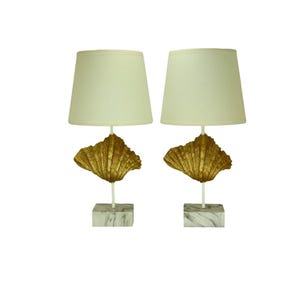 Può includere: Coppia di lampade da tavolo decorative. Ogni lampada presenta un paralume color crema, un design a conchiglia dorata, uno stelo bianco e una base marmorizzata. Le lampade sono progettate per uso interno.
