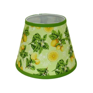 May include: A tapered lampshade with a green rim and a light green background. The lampshade is decorated with a pattern of lemons, green leaves, and white flowers. The lemons are yellow, and the leaves are a deep green.