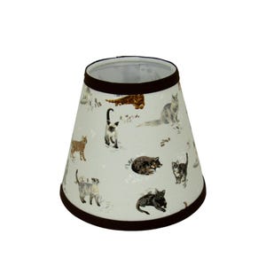 May include: A light beige lampshade with a brown trim features a repeating pattern of cats in various poses. The cats are depicted in shades of brown, gray, and white.