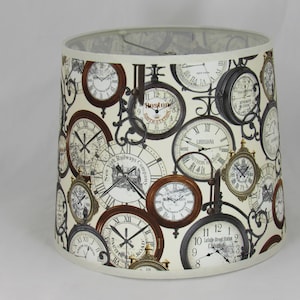 Pode incluir: Um abajur branco com um padrão de relógios antigos. Os relógios têm numerais romanos e são de vários estilos. Os relógios têm detalhes pretos, marrons e brancos.