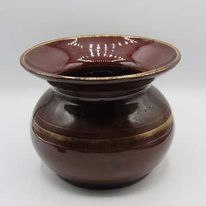 May include: A dark red ceramic vase with a wide, flared opening and a rounded body. The vase has two thin gold bands around the middle.