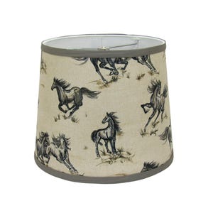 Op de afbeelding: Cilindrische lampenkap met een herhaald patroon van rennende paarden in grijstinten en bruin op een beige achtergrond. De boven- en onderranden zijn afgewerkt met een grijze rand. De lampenkap is ontworpen om over een standaard lampvoet te passen.