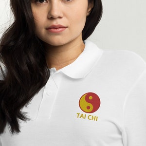 May include: White polo shirt with a collar and short sleeves. The shirt features an embroidered Tai Chi symbol in red and yellow, with the words "TAI CHI" below it in gold. The shirt is made of a soft material.