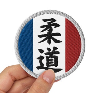 Peut inclure: Un écusson rond brodé aux couleurs du drapeau français : bleu, blanc et rouge. Le patch porte les caractères japonais pour le Judo en noir. Le patch est tenu par une main.