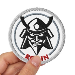 Könnte beinhalten: Ein runder weißer Patch mit einem gestickten Schwarzweißbild eines Samurai-Kriegergesichts mit dem roten gestickten Wort "ROKIN" unter dem Bild.