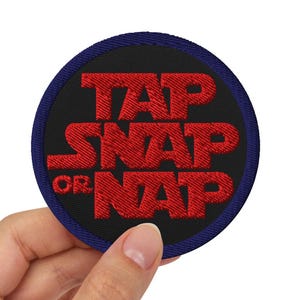 May include: A circular embroidered patch featuring the text "TAP SNAP OR NAP" in red, set against a black backdrop, and encircled by a navy blue border. The patch is held by a hand.