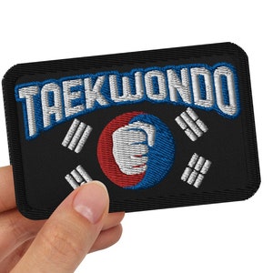 May include: A black embroidered patch with the word "Taekwondo" in white lettering and a red and blue fist in the center. The fist is surrounded by a white circle and a black background with white stripes.