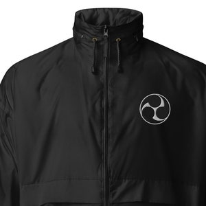 Op de afbeelding: Een zwarte windbreaker-jas met een ritssluiting en een klein geborduurd symbool op de linkerborst. Het symbool is een witte gestileerde ster met drie punten.