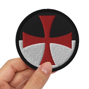Op de afbeelding: Een ronde, zwarte en witte geborduurde patch met een rood kruis in het midden. De patch wordt in een hand gehouden.