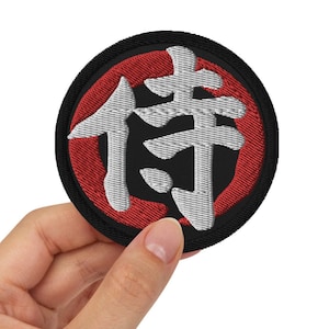 Könnte beinhalten: Ein runder schwarzer und roter Patch mit weißen gestickten japanischen Schriftzeichen, die "侍" (Samurai) lauten.