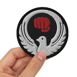 May include: A black and white embroidered patch featuring a red fist and a white bird in the centre. The patch is circular and has a black border.