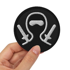 Peut inclure: Un écusson circulaire noir avec un motif brodé blanc représentant une personne portant un masque et tenant deux épées.