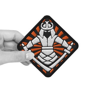 Könnte beinhalten: Ein gestickter schwarzer und orangefarbener Aufnäher mit einem weißen Samurai-Krieger mit Helm und Schwert. Der Krieger steht in einem strahlenden Sonnenstrahlenmuster.
