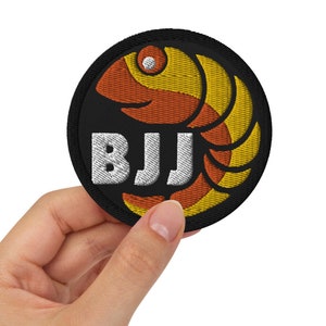 Puede incluir: Un parche redondo negro con un diseño de pez bordado en amarillo y naranja y el texto "BJJ" en blanco.