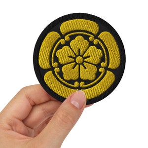 Könnte beinhalten: Ein gestickter Aufnäher in Schwarz und Gold mit einem stilisierten Blumendesign. Der Aufnäher ist rund und hat einen schwarzen Rand.