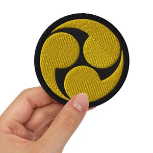 Puede incluir: Un parche bordado negro y amarillo con un símbolo de tres partes. El símbolo es un diseño estilizado de tres formas curvas superpuestas.