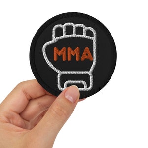 Peut inclure: Un écusson brodé noir et blanc avec un poing blanc et le mot "MMA" en orange.