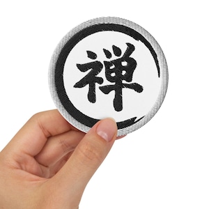 Pode incluir: Um remendo branco redondo com um círculo preto e a palavra japonesa "Zen" escrita em preto dentro do círculo.