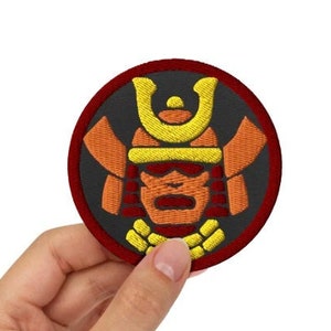 Könnte beinhalten: Ein runder gestickter Aufnäher mit rotem Rand und schwarzem Hintergrund. Der Aufnäher zeigt einen gelben und orangefarbenen Samurai-Krieger, der einen Helm mit einem gelben Wappen trägt.