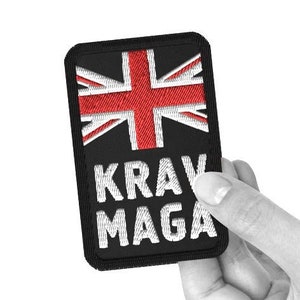 Puede incluir: Parche rectangular negro con la bandera de la Union Jack roja y blanca en la parte superior. Las palabras "KRAV MAGA" están bordadas en blanco debajo. El parche se muestra sobre un fondo blanco.