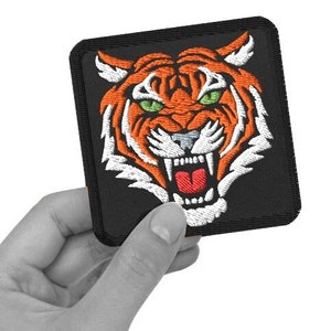 Puede incluir: Un parche bordado negro con la cara de un tigre. El tigre tiene rayas naranjas y blancas y una boca roja abierta.