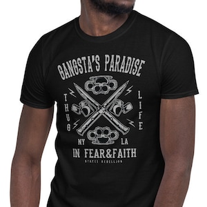 May include: Black t-shirt with a white graphic design. The graphic features a pair of crossed knives and brass knuckles with the text "Gangsta's Paradise", "Thug Life", "NY", "LA", "In Fear & Faith", and "First Rebellion".