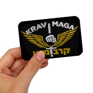 Puede incluir: Un parche bordado negro con alas doradas y una espada. El texto "KRAV MAGA" está escrito en blanco sobre las alas y el texto "לוחמה" está escrito en blanco debajo de las alas.