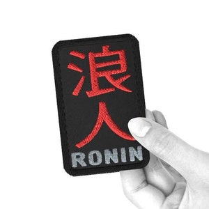 Könnte beinhalten: Schwarzer gestickter Aufnäher mit roten japanischen Schriftzeichen und dem Wort "RONIN" in grauer Schrift.