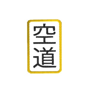 Peut inclure: Écusson brodé avec les caractères japonais pour "Karate-Do" en noir sur fond blanc. L'écusson est rectangulaire avec un bord doré. Le texte est orienté verticalement.
