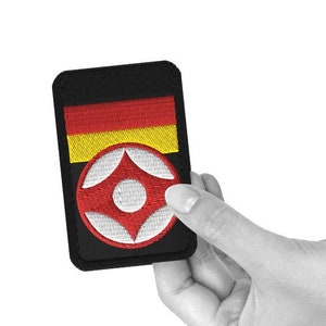 May include: A black embroidered patch with a red, white, and yellow design. The patch features a circle with a red center and white spokes.