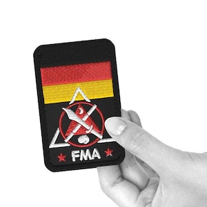 Pode incluir: Etiqueta bordada preta com uma bandeira alemã vermelha, amarela e preta, um triângulo vermelho com espadas cruzadas e o texto "FMA" em branco.