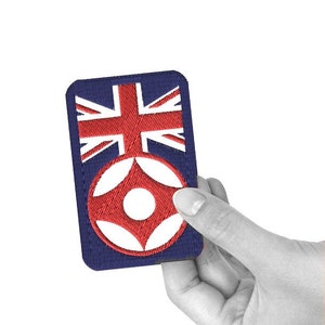 May include: A navy blue embroidered patch featuring a red and white design over a red and white Union Jack. The patch is rectangular with rounded corners. The patch is held in a hand.