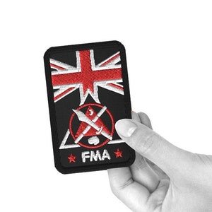 Peut inclure: Écusson brodé noir avec un drapeau Union Jack rouge, un cercle rouge avec des épées croisées, un triangle blanc et le texte "FMA" avec deux étoiles blanches.