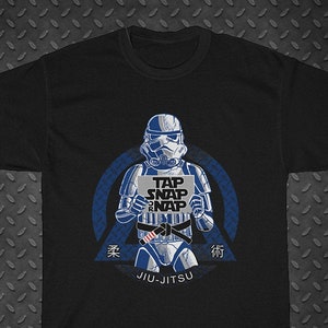 May include: Black t-shirt featuring a blue and white illustration of a Star Wars Stormtrooper wearing a blue belt and holding a sign that says "Tap Snap Nap" with the text "Jiu-Jitsu" below the illustration.