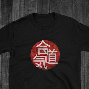 Może przedstawiać: Czarny T-shirt z czerwonym kółkiem i białymi japońskimi znakami, które brzmią "合氣道" (Aikido).