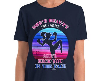 May include: A navy blue youth t-shirt with a graphic of a woman kicking in a sunset-colored circle. The text reads "She's Beauty She's Grace She'll Kick You In The Face".