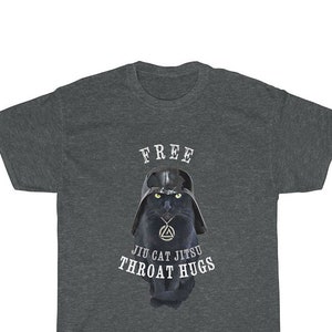 May include: A dark heather grey t-shirt featuring a black cat wearing a Darth Vader helmet and a pendant. The text on the shirt reads "FREE JIU CAT JITSUI THROAT HUGS".