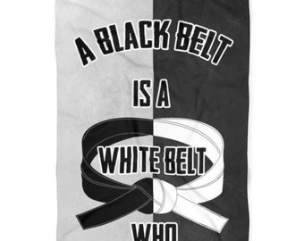 May include: A martial arts-themed towel, split vertically into black and white sections. The text reads "A Black Belt Is A White Belt Who Never Quit." Features a black and white belt design, with gold stars.