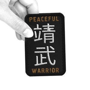 May include: Black patch with white text that reads "Peaceful Warrior" in English and Chinese characters. The patch is held in a hand.