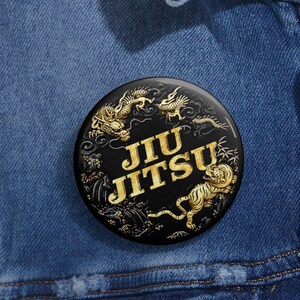May include: A black button with the words "Jiu Jitsu" in gold lettering. The button features gold illustrations of a dragon and a tiger. The button is pinned to a blue denim jacket.