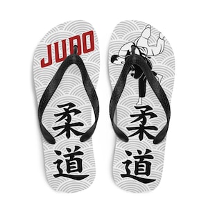 Peut inclure: Paire de tongs blanches avec une sangle noire. Une tong présente une silhouette noire d'un lancer de judo, tandis que l'autre porte le mot "JUDO" en lettres rouges. Les deux tongs affichent les caractères japonais pour "Judo" en noir.