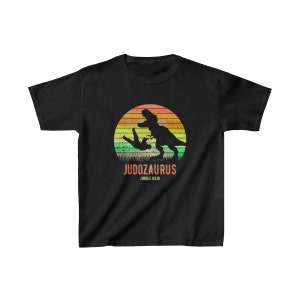 May include: Black t-shirt with a retro sunset graphic of a dinosaur doing a judo throw on a person. The text "Judozaurus Jungle Dojo" is below the graphic.
