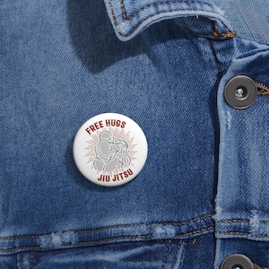 May include: White button with a red and white graphic of two people in a hug with the text "Free Hugs Jiu Jitsu" around the image.