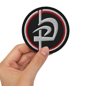 May include: Round black embroidered patch with a white and red design. The patch features a stylized symbol in white, outlined with a red border, and a black background. The patch is held in a hand.