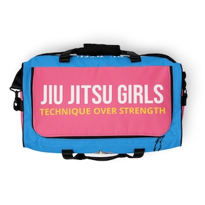 Op de afbeelding: Een roze en blauwe duffelbag met de tekst "Jiu Jitsu Girls" en "Technique Over Strength" in witte en gele letters.