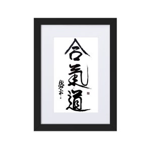 May include: Framed artwork featuring Japanese calligraphy in black ink on white paper. The calligraphy reads "Aikido" in stylized characters. The artwork is set in a black frame with a white mat.