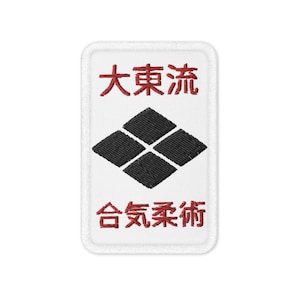 Op de afbeelding: Witte geborduurde patch met rode tekst die "大東流 合気柔術" luidt en een zwarte ruit in het midden.