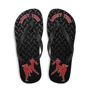 May include: Pair of black flip flops with a black and grey diamond pattern. The flip flops have the words "Muay Thai" in red on the straps and a red silhouette of two women practicing Muay Thai.